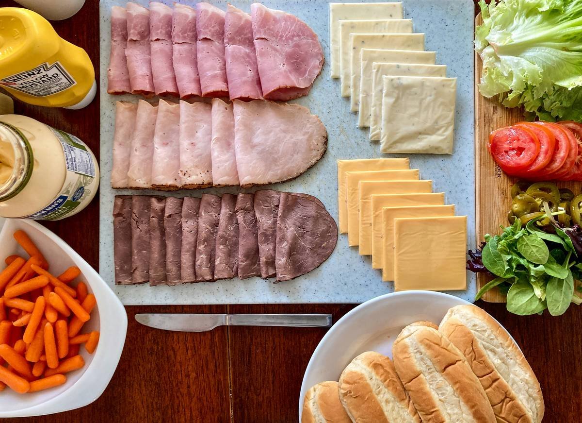 A delicious lunch spread of deli meats, cheese, tomato, lettuce, carrots, mustard, mayo, other condiments, jalapenos, spinach, hoagie buns ready for a family lunch of subway style sub sandwiches.